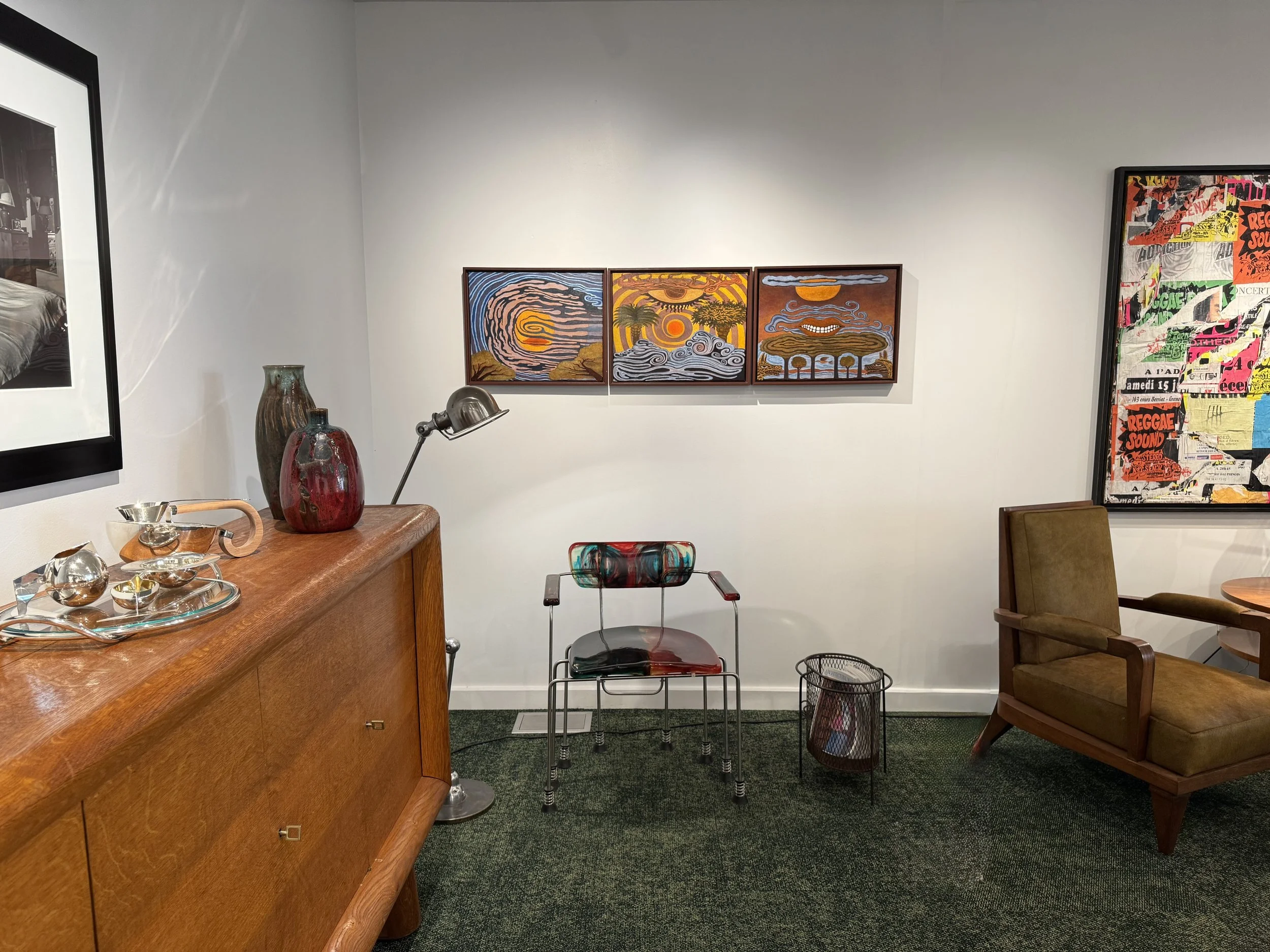 An interior room with a wooden sideboard displaying various glassware, decorative vases, and a small bowl. Behind the sideboard are three framed colorful paintings of abstract landscapes hanging on a plain white wall. To the right, there is a brown upholstered armchair and a mustard yellow chair. In the middle of the room, there is a transparent chair made of colorful glass or plastic with a metal frame. To the left, part of a black picture frame with a photo is visible, and to the right, part of a collage-style artwork with colorful text and images. There is also a metallic adjustable desk lamp, a small round table, and a wire wastebasket.
