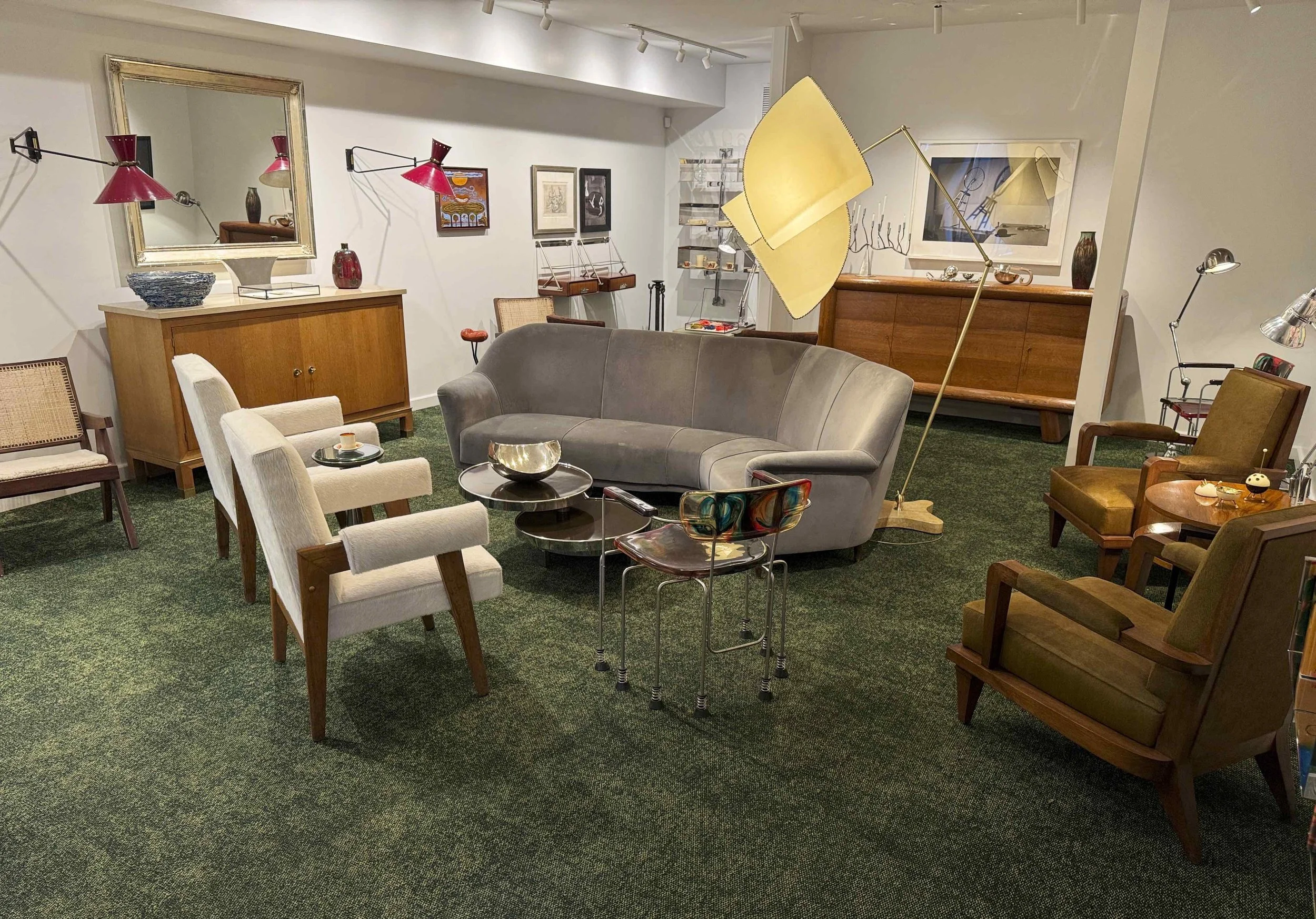 A cozy living room with various vintage and modern furniture including a gray curved sofa, white armchairs, and wooden chairs. Decorative lamps, artwork, and vases adorn the room, along with a large mirror and a green carpet.