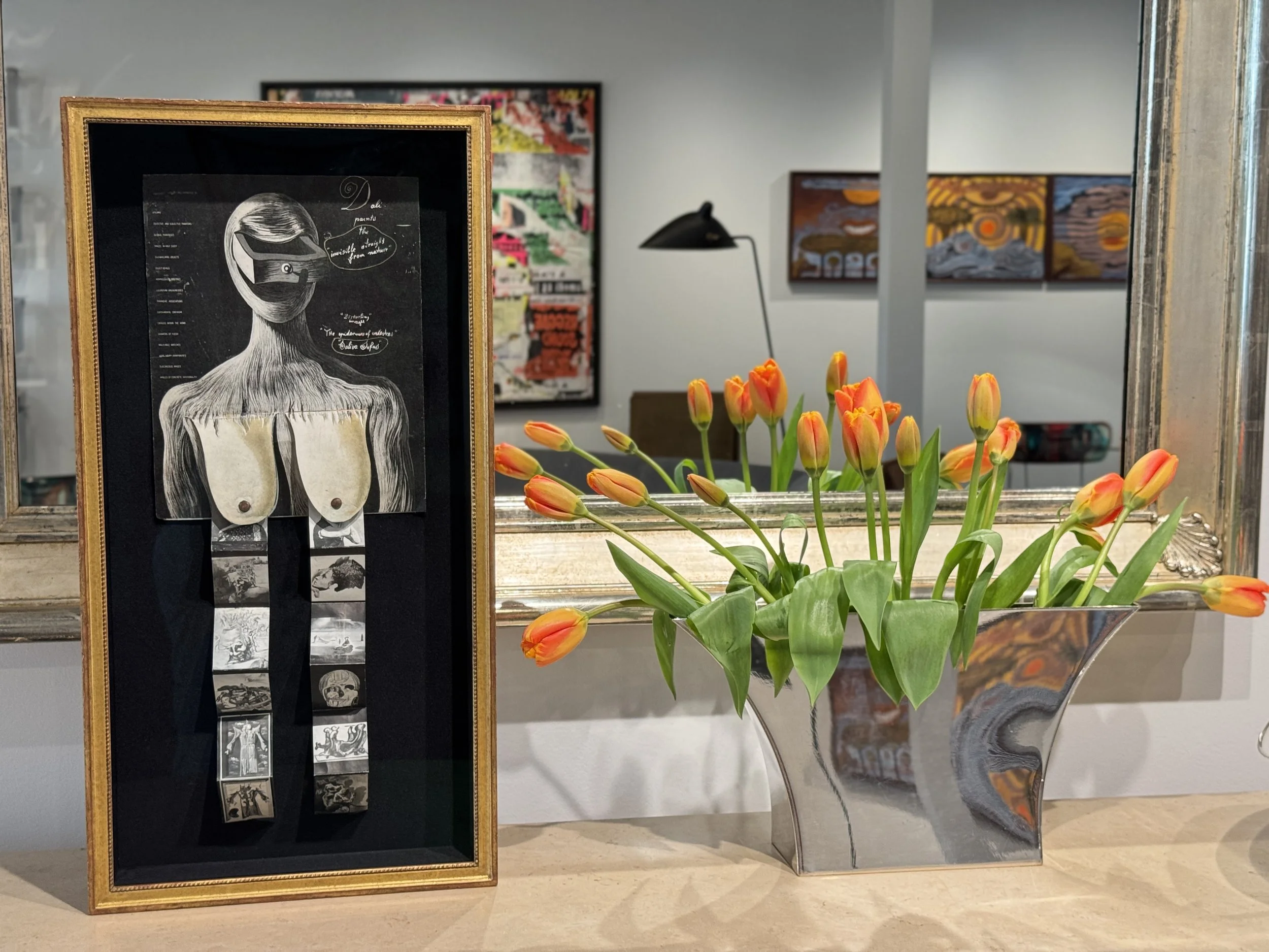 Framed artwork depicting a stylized female figure with short hair, a mask over her eyes, and exposed shoulders, surrounded by smaller scratched or drawn panels, beside a vase of orange tulips, in an art gallery.