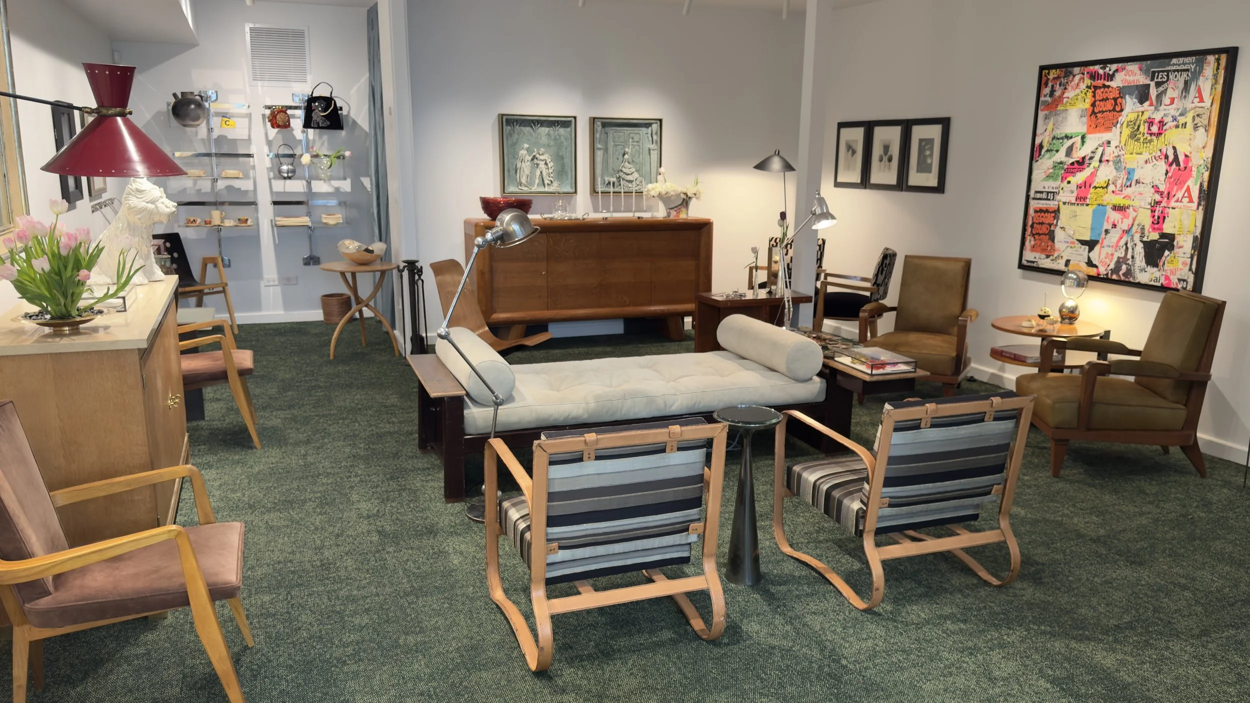 A vintage-style living room with various chairs, a white tufted sofa, a wooden sideboard, and framed artwork on the walls. There are decorative items including a red lamp, flowers, and sculptures, with a green carpet covering the floor.