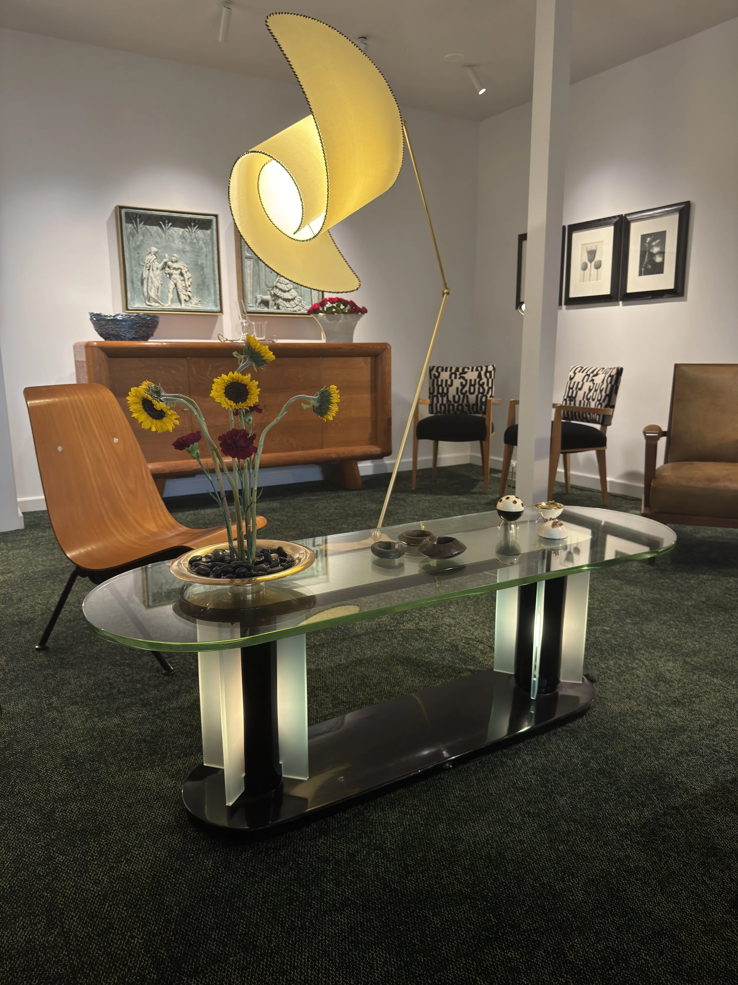 Modern living room features a glass coffee table with sunflower and red flowers, a wooden console behind it, a yellow arc-shaped standing lamp, framed artwork on the walls, and various chairs including a wooden chair and black and patterned upholstered chairs.