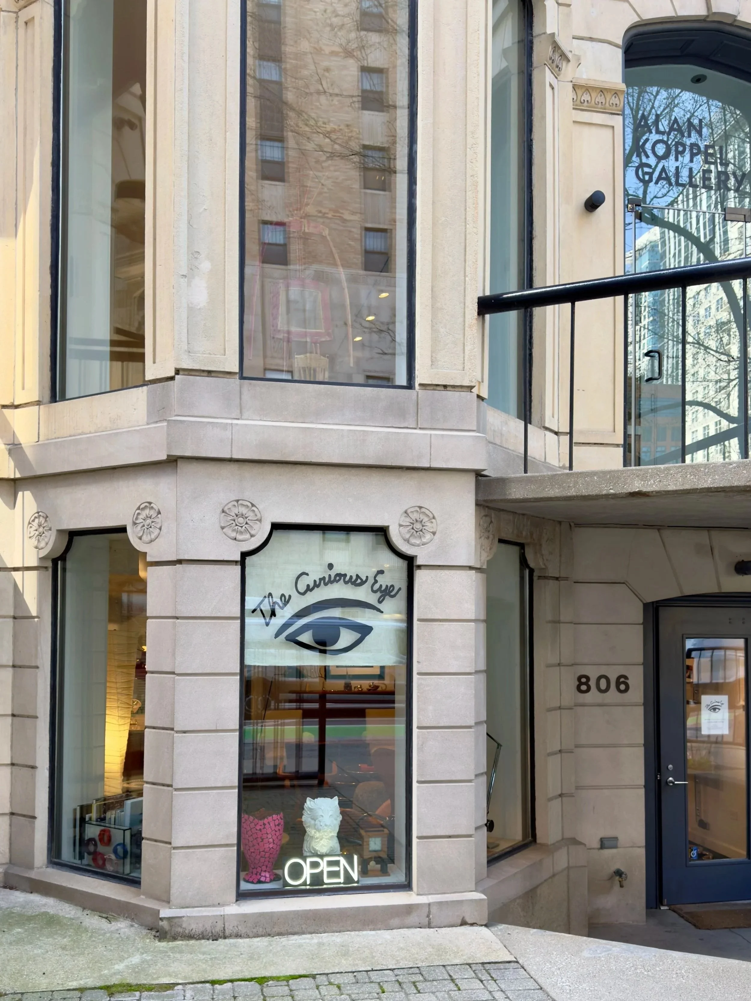 The exterior of an art gallery called Alan Koppel Gallery located at 806. The storefront has large glass windows with decorative stone framing, showcasing art pieces and decorative objects inside. There is a neon "OPEN" sign in the window, and the gallery's logo featuring an eye with the words "The Curious Eye" is displayed. The entrance is to the right, with a glass door and a black railing leading to it.