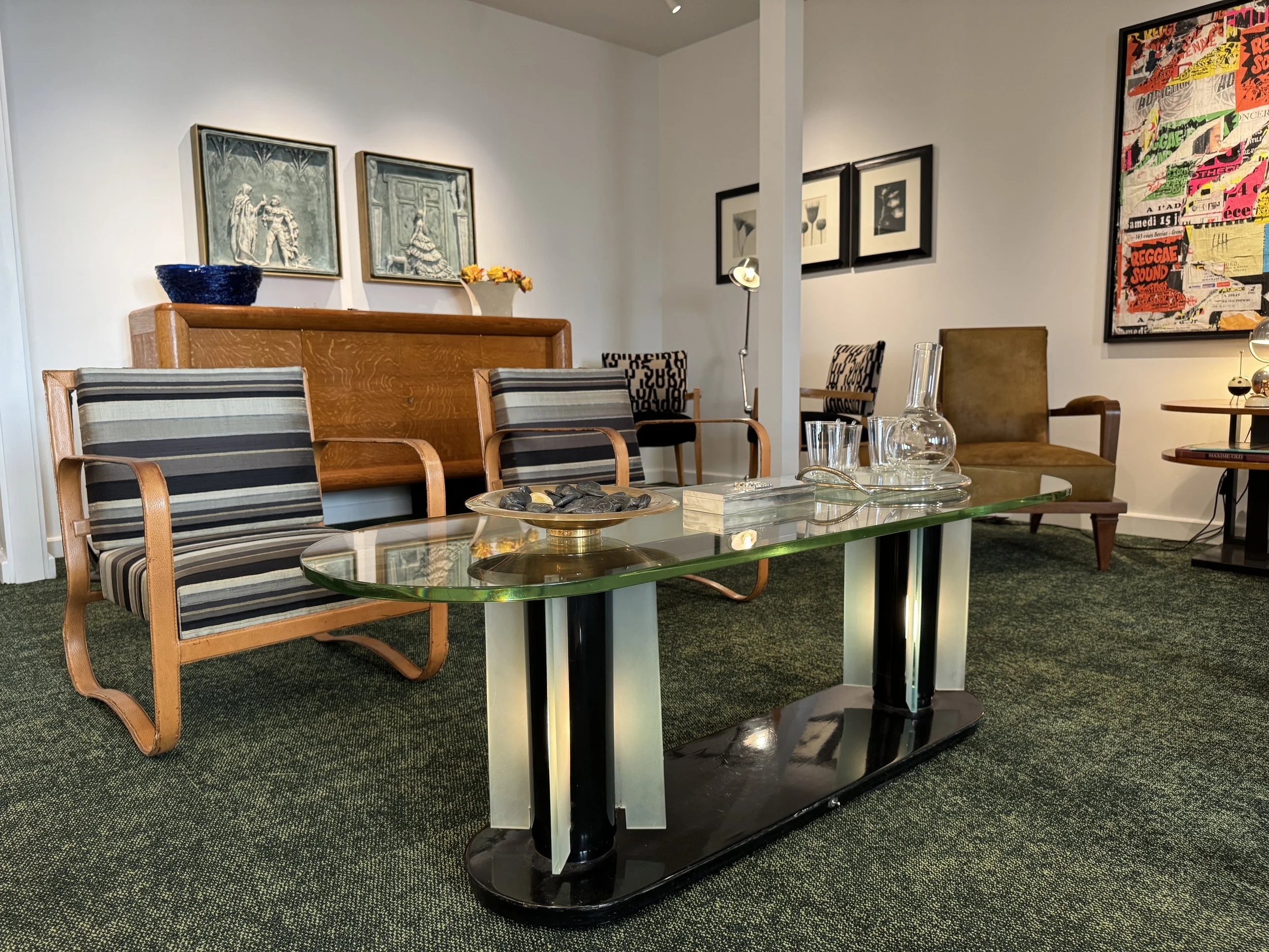 Living room with wooden and upholstered chairs, a glass-topped coffee table with decorative stones, a modern black base lamp, framed artwork on the walls, and a collaged poster with bright colors.