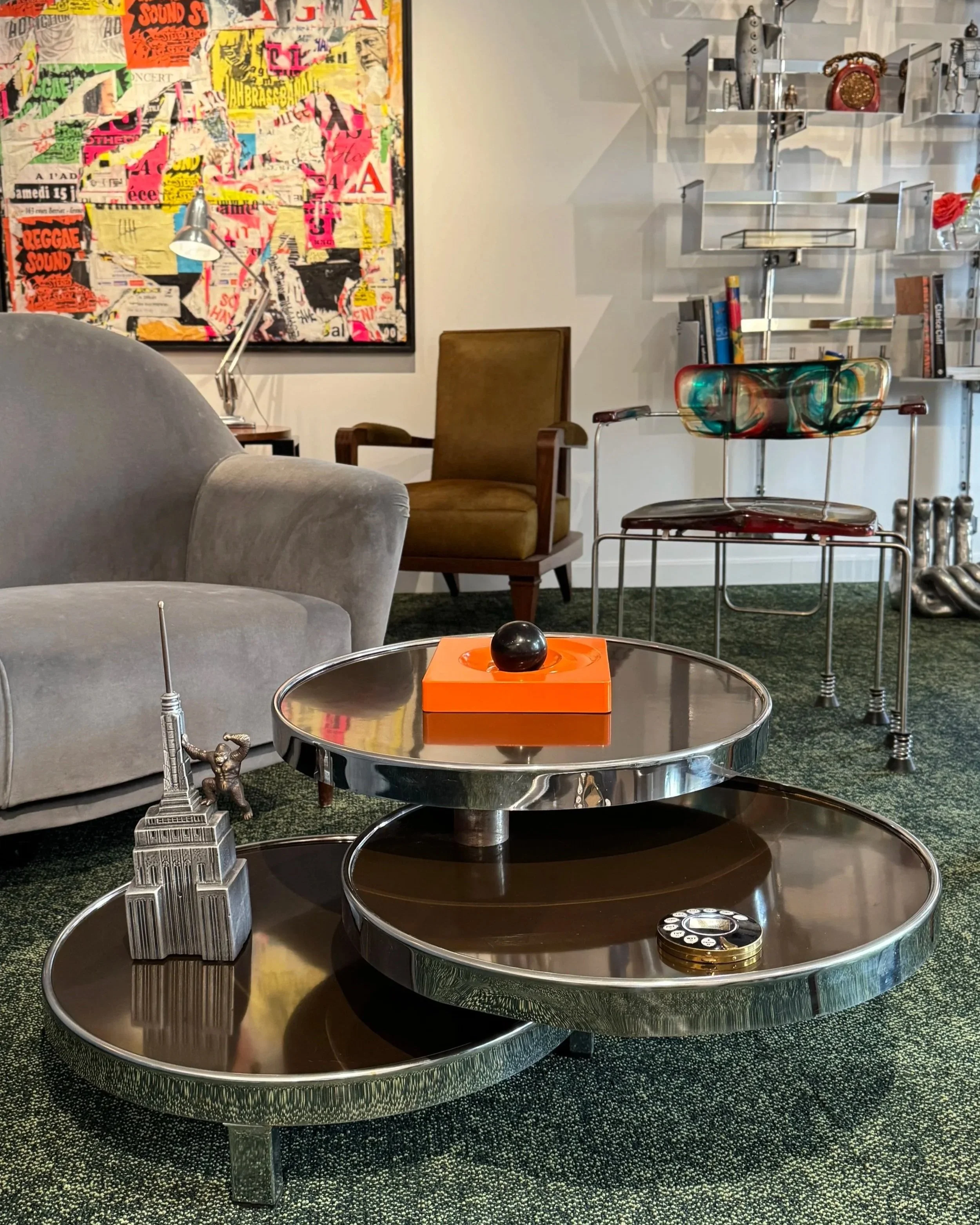 Living room with modern furniture, a large colorful abstract painting, a metal side table with a decorative miniature Eiffel Tower and a man sculpture, a brown armchair, a tall floor lamp, a tiered glass wall shelf with decorative objects, and a coffee table with a tissue box, a black sphere, and a small round object.