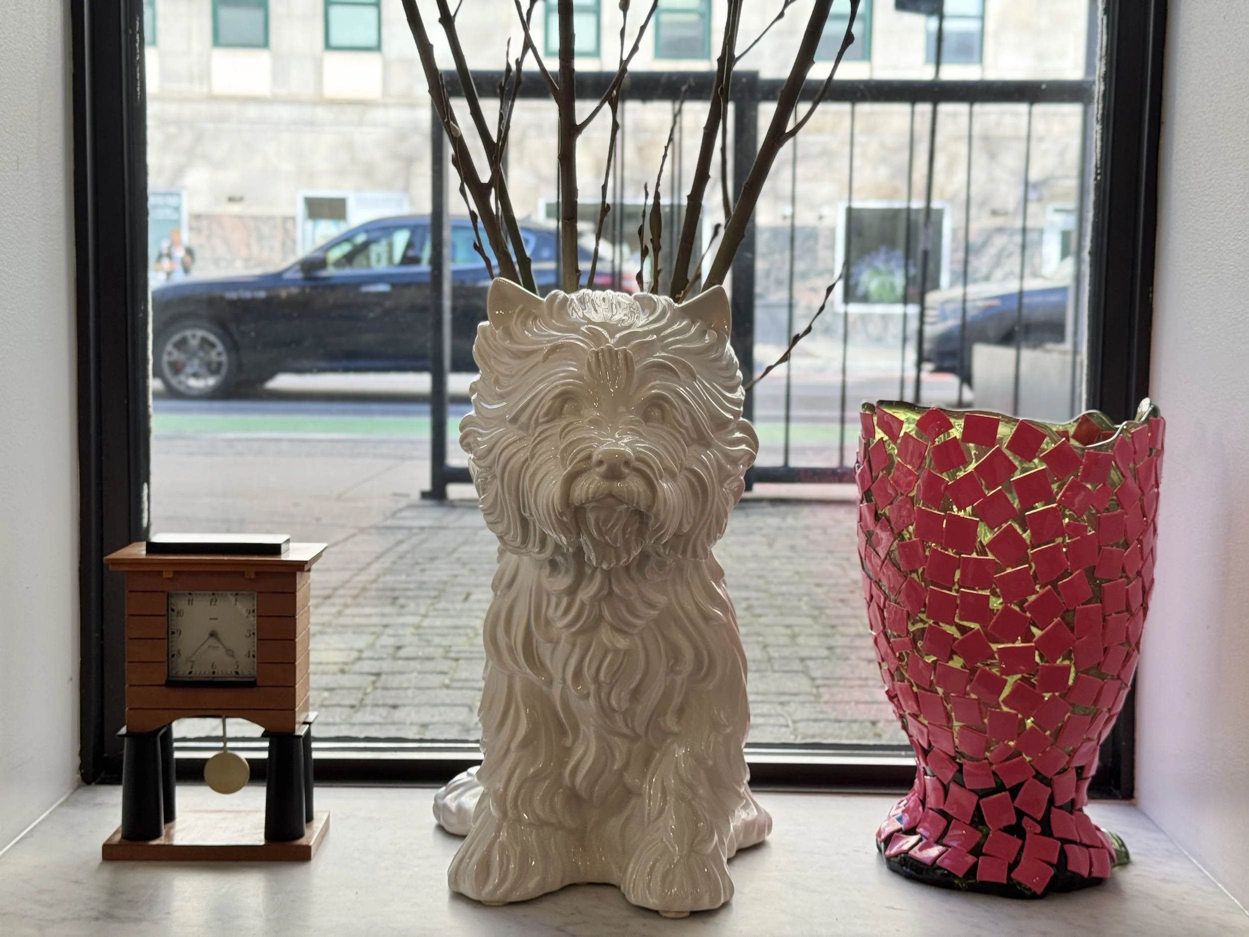 A ceramic lion head vase filled with tree branches sitting on a windowsill. To its right is a tall pink mosaic vase and to its left is a wooden clock on a small table, with a parking lot and a black car visible outside the window.