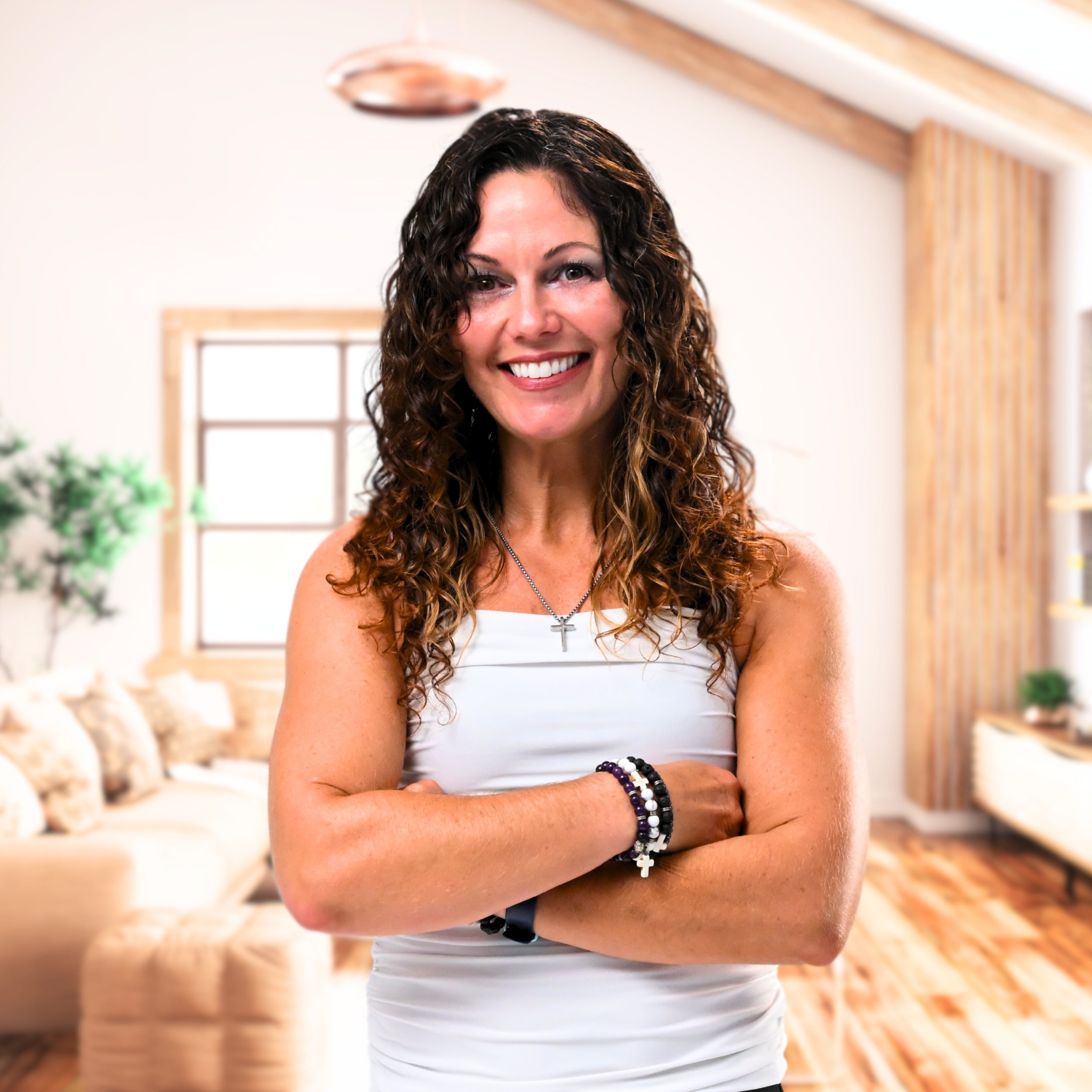 smiling woman with curly brown hair wearing a white tank top and cross necklace, standing with arms crossed in a cozy living room with beige walls, wooden accents, and a couch.