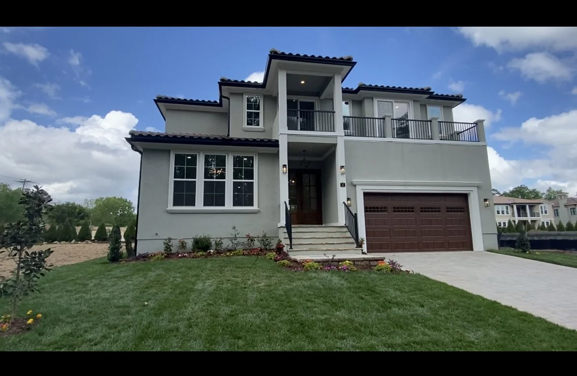 Modern two-story house with a front porch, large windows, and a two-car garage, surrounded by a well-maintained lawn and small garden, under a blue sky with clouds.