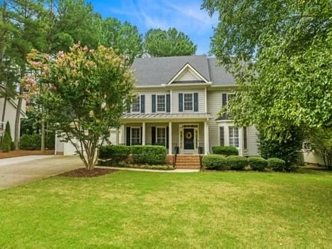 Front view of a two-story house with a manicured lawn, flowering tree, and green shrubs, under a blue sky with green trees in the background.