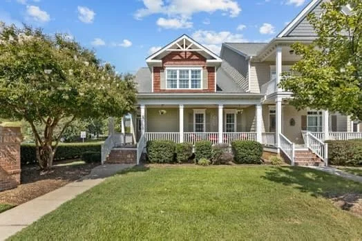 Front view of a suburban house with a large front porch, neatly trimmed bushes, a well-maintained lawn, and trees, with a blue sky and some clouds in the background.