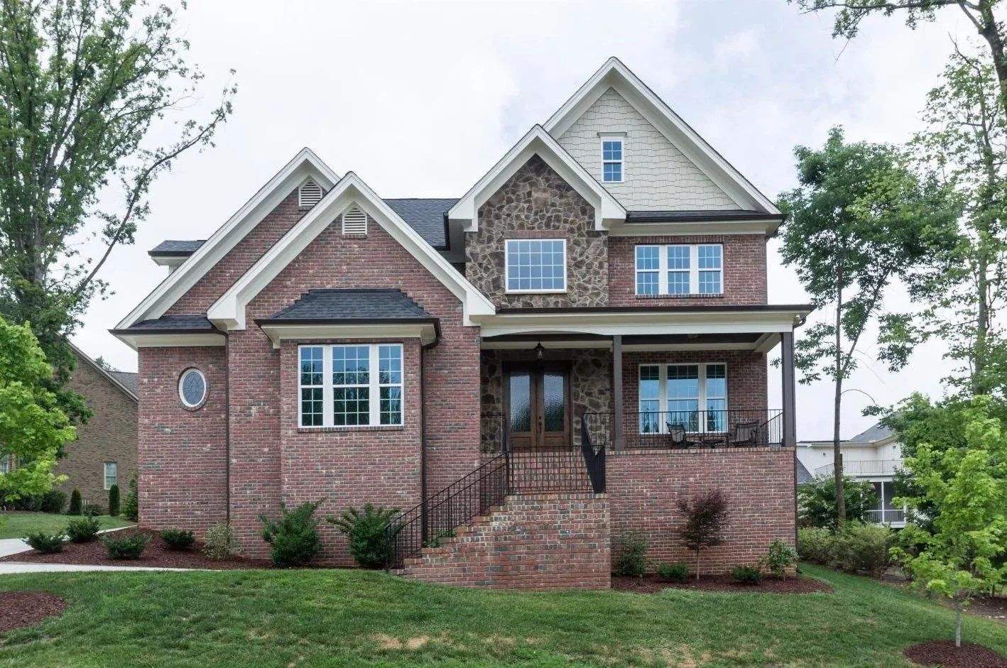 A large two-story house with a brick exterior, stone accents, and multiple windows, featuring a front porch and stairs leading up to the entrance, surrounded by greenery and trees.