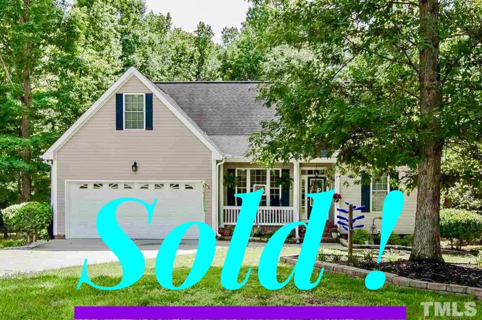 A house with beige siding, a two-car garage, front porch, surrounded by trees and shrubs, with a large 'Sold!' text overlay in light blue.