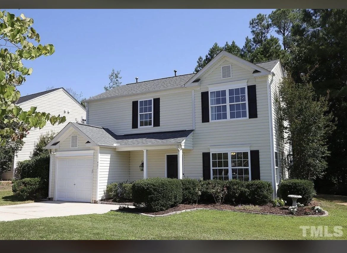 White two-story house with black shutters, attached garage, front porch with a small roof, well-maintained lawn, shrubs, and trees in the background on a clear, sunny day.