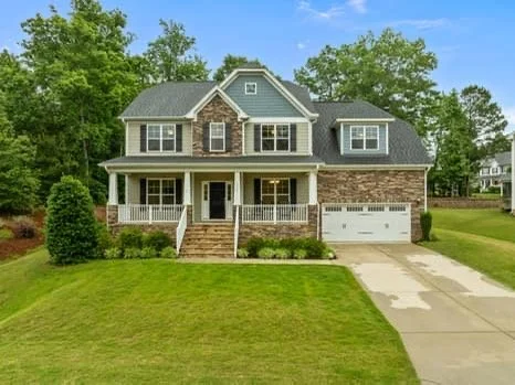 A two-story suburban house with gray roofing, brick and siding exterior, front porch, and attached garage, surrounded by a well-maintained lawn and trees.