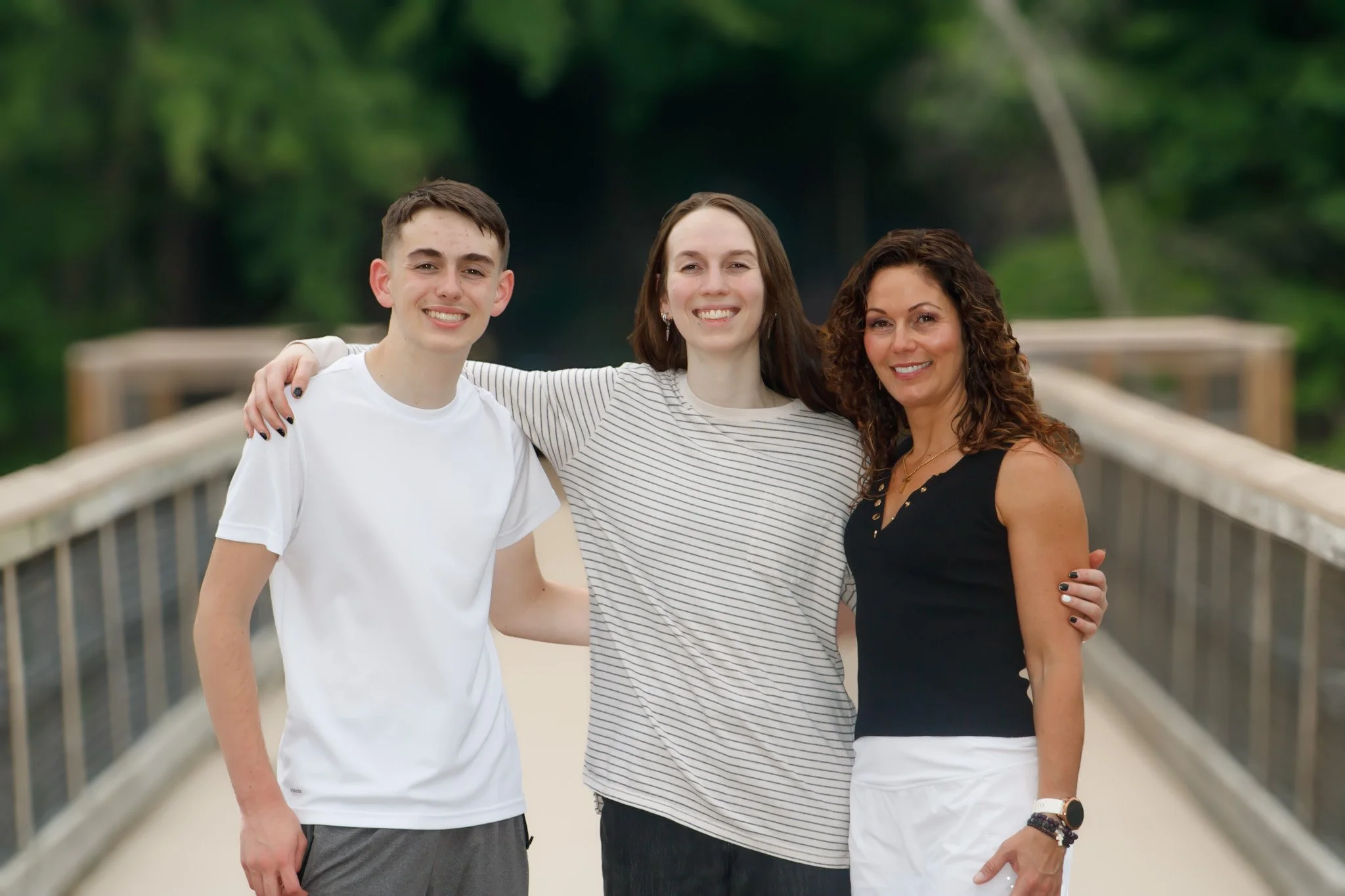 Three people standing close together on a bridge, smiling at the camera. The group includes a young man on the left, a woman in the middle, and a woman on the right with curly hair. They are outdoors with green trees in the background.