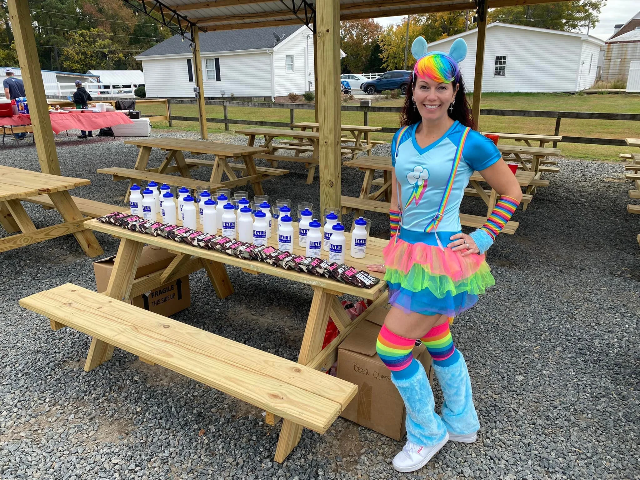 Woman dressed in rainbow-colored unicorn costume standing next to a table with water bottles and snack bars at an outdoor event.