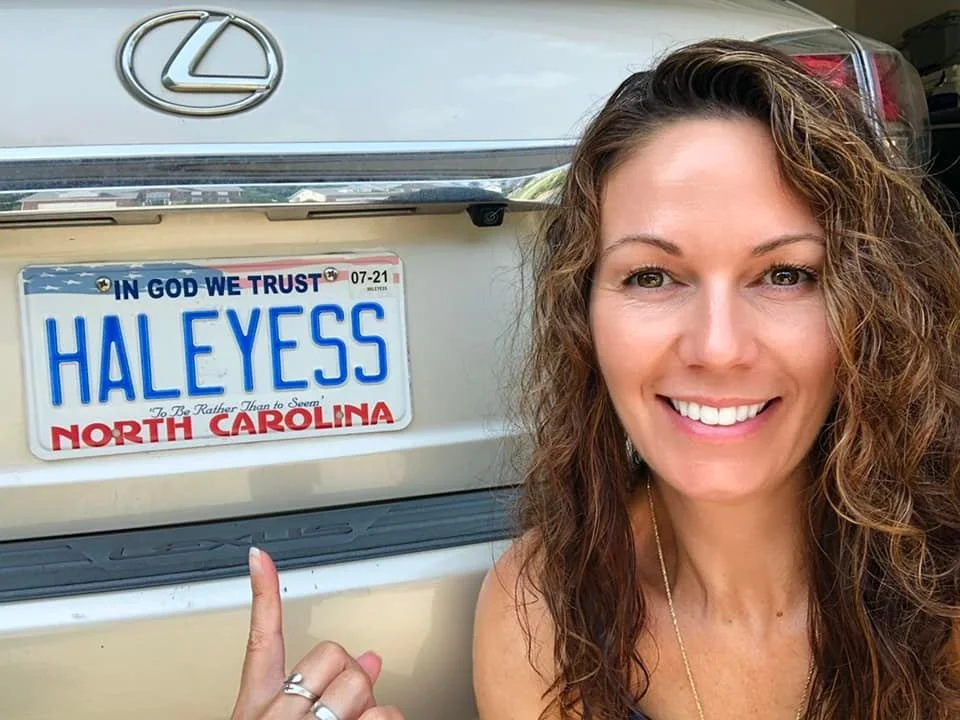 Woman with curly hair smiling and pointing at a North Carolina license plate on a beige Lexus car.