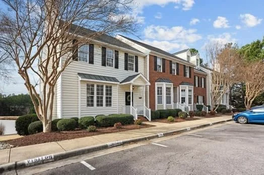 A row of townhouses with white and red brick facades, a parking lot in front, and a blue car parked nearby under a partly cloudy sky.