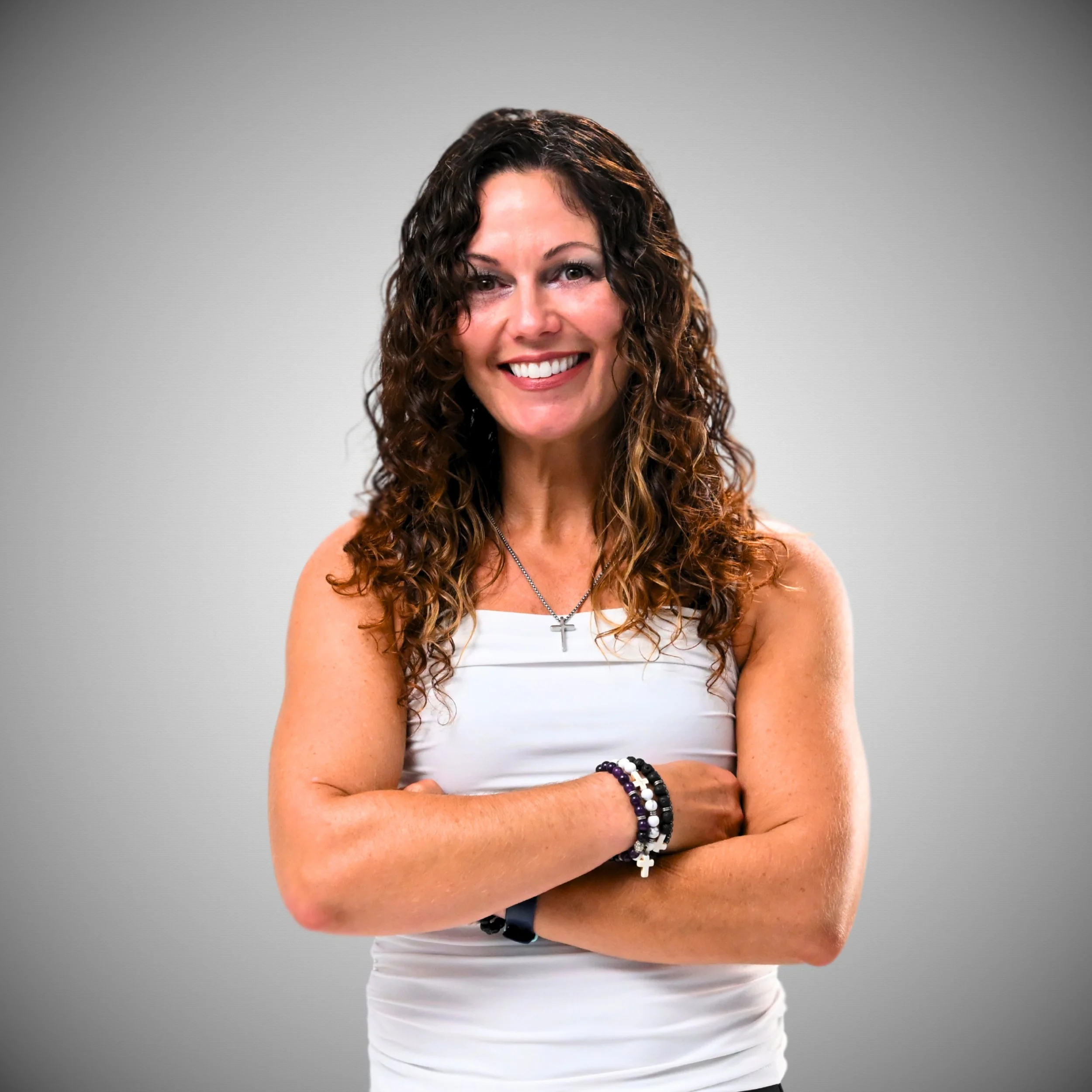A woman with curly brown hair smiling, wearing a white sleeveless top, cross necklace, and bracelets on her left wrist, standing against a plain gray background.