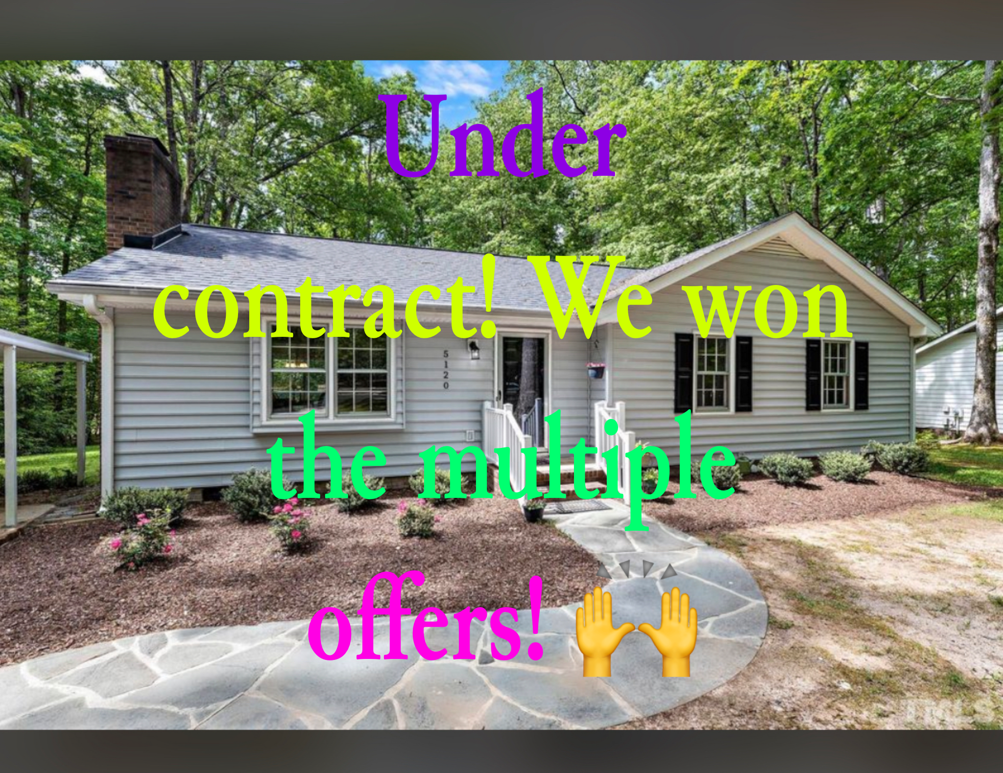 A house with a well-maintained front yard featuring a stone pathway, small bushes, and a forested background with trees. Overlaid text reads: 'Under contract! We won the multiple offers!' with colorful letters and a hand-raising emoji.