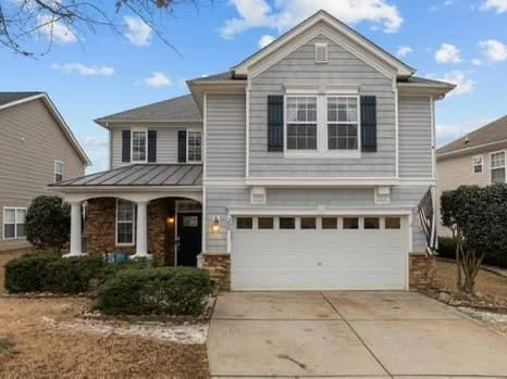 A two-story suburban house with a white garage door and blue shutters, front porch, and a driveway leading to the street.