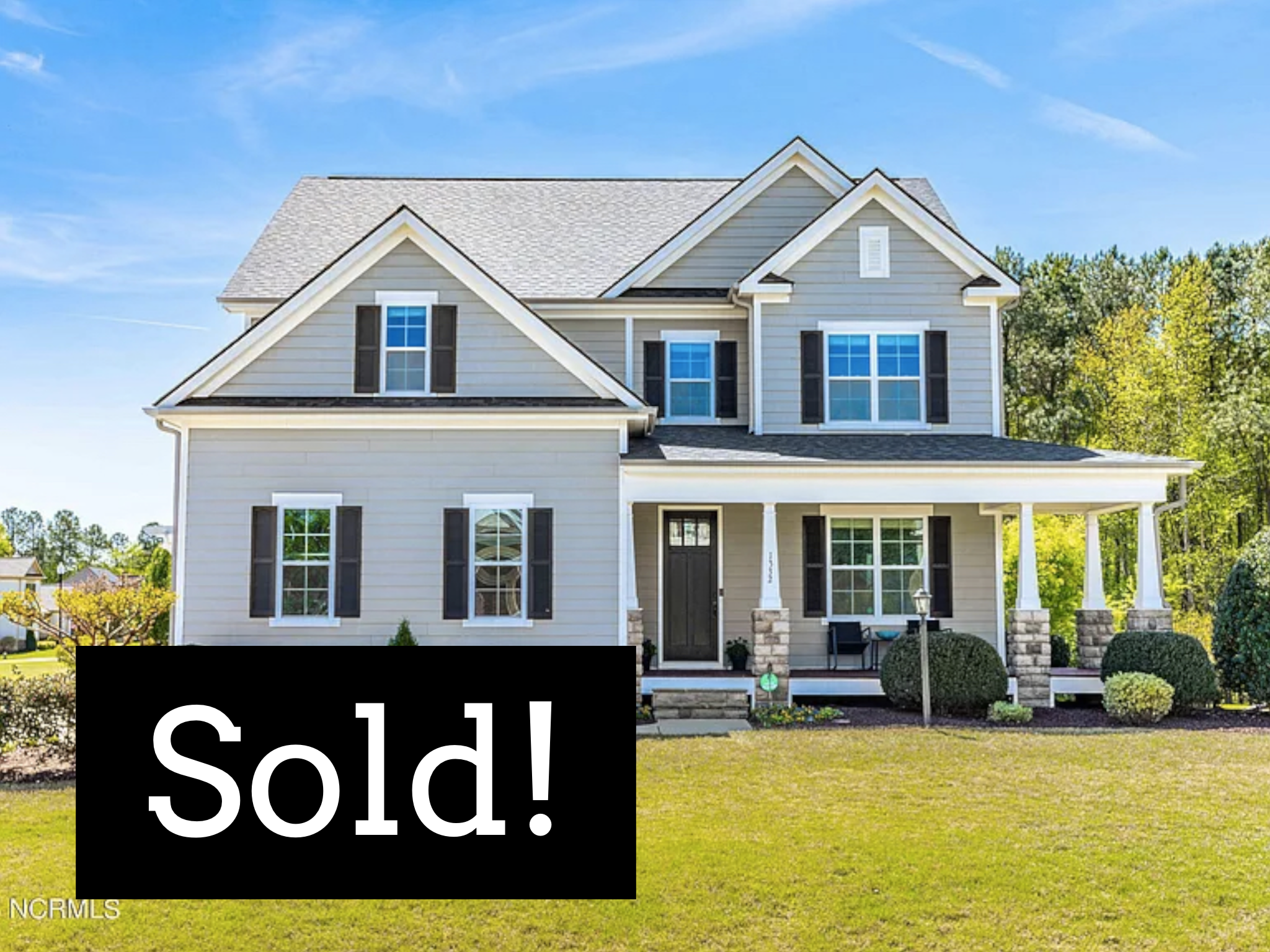 A large two-story suburban house with a front porch, black front door, multiple windows with black shutters, and a well-maintained lawn. A black sign with white text reads 'Sold!'.