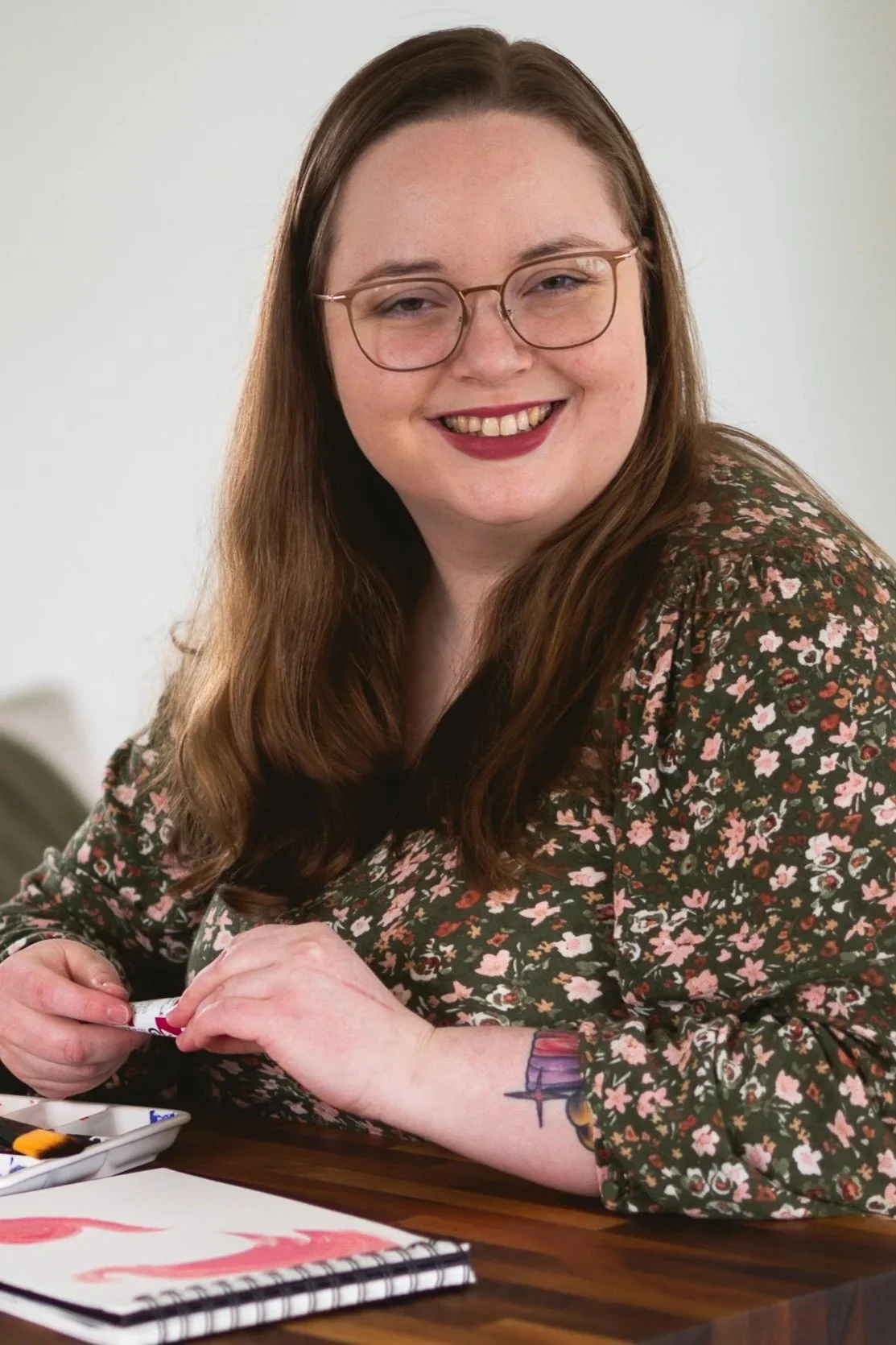 A therapist with long brown hair, glasses, and a floral shirt is smiling and sitting at a table, holding a paintbrush and working on a painting.