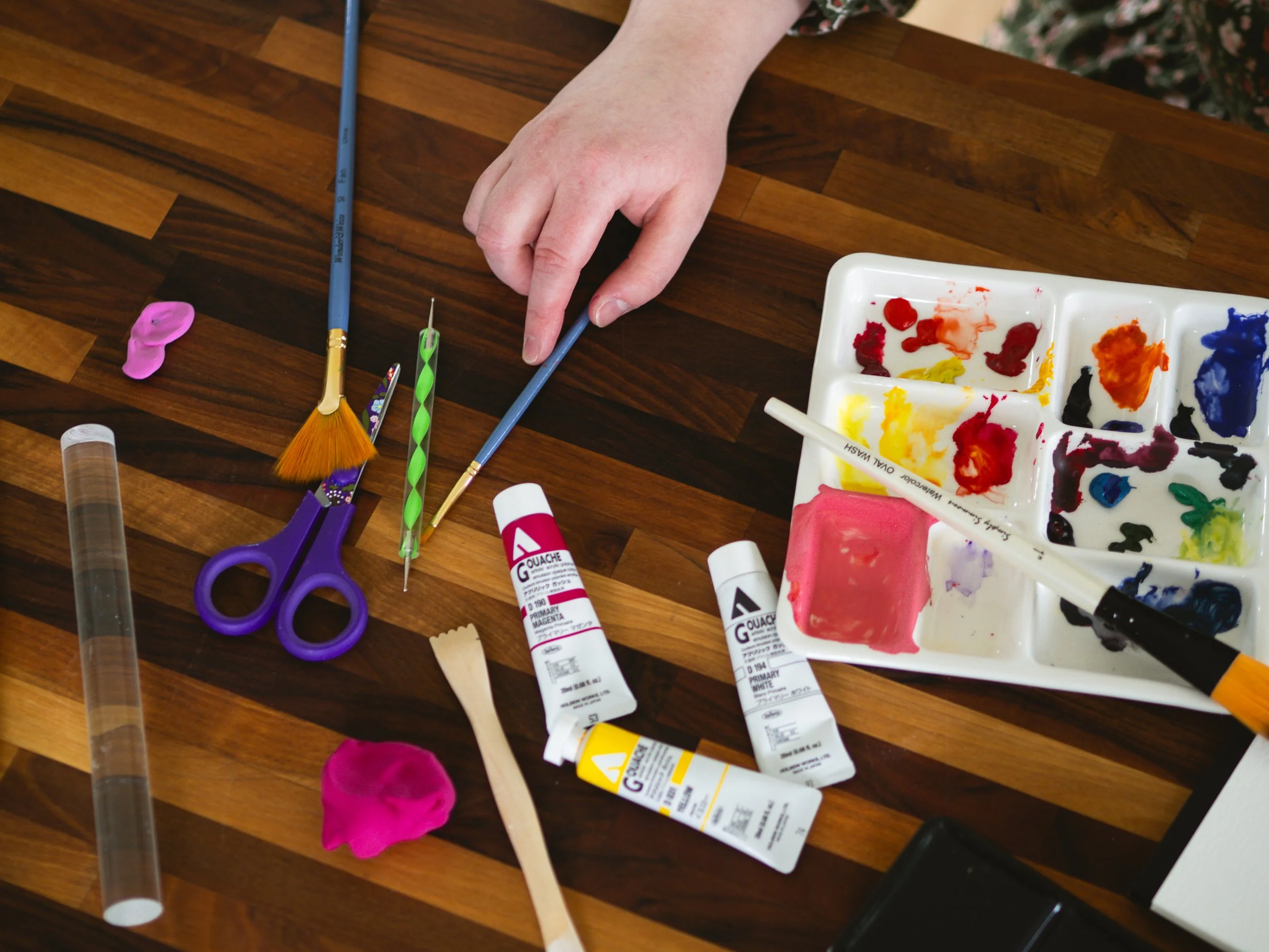 A person's hand arranging paintbrushes and art supplies on a wooden surface, with paint tubes, a paint palette, scissors, and paint blobs visible.