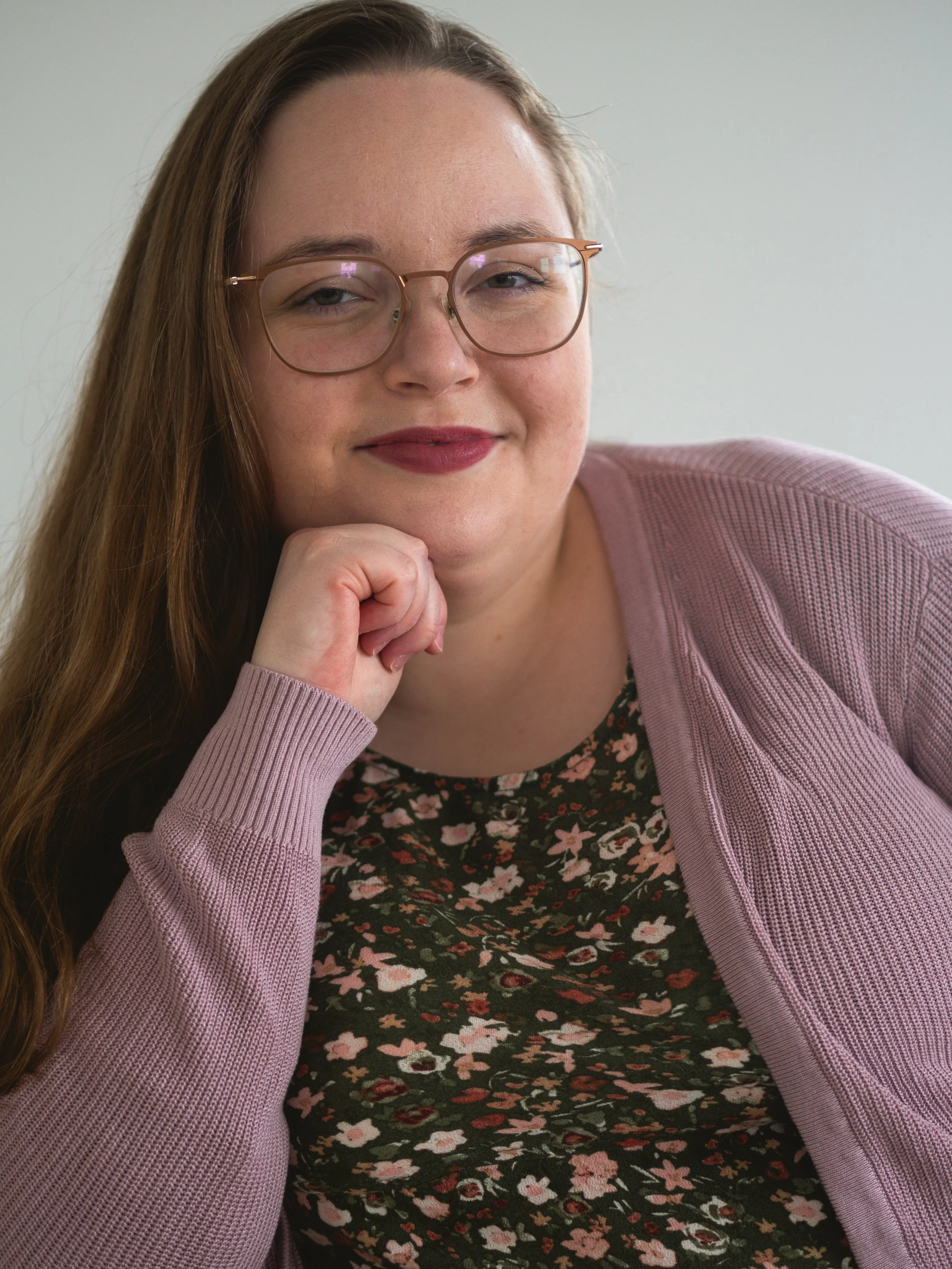 Therapist with long brown hair, wearing glasses and a floral dress, resting her chin on her hand and smiling softly.