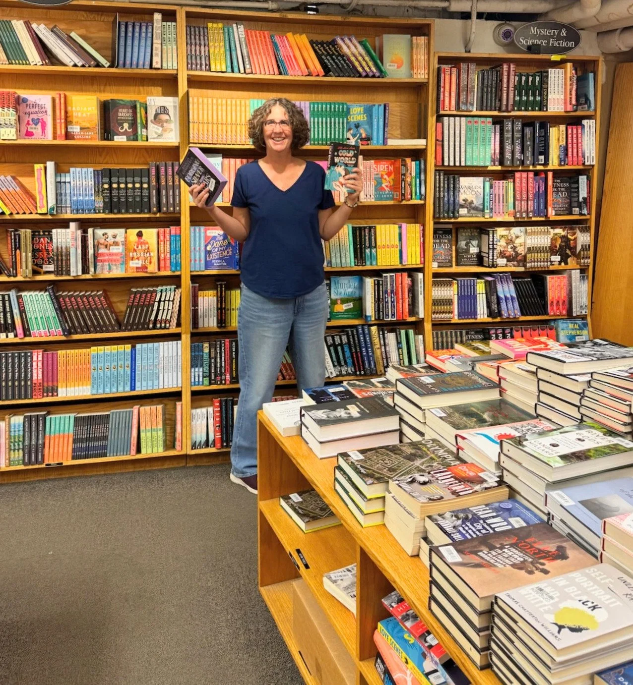 A woman with curly hair wearing a blue shirt and jeans standing in a bookstore, smiling and holding books in both hands. The background features bookshelves filled with colorful books.