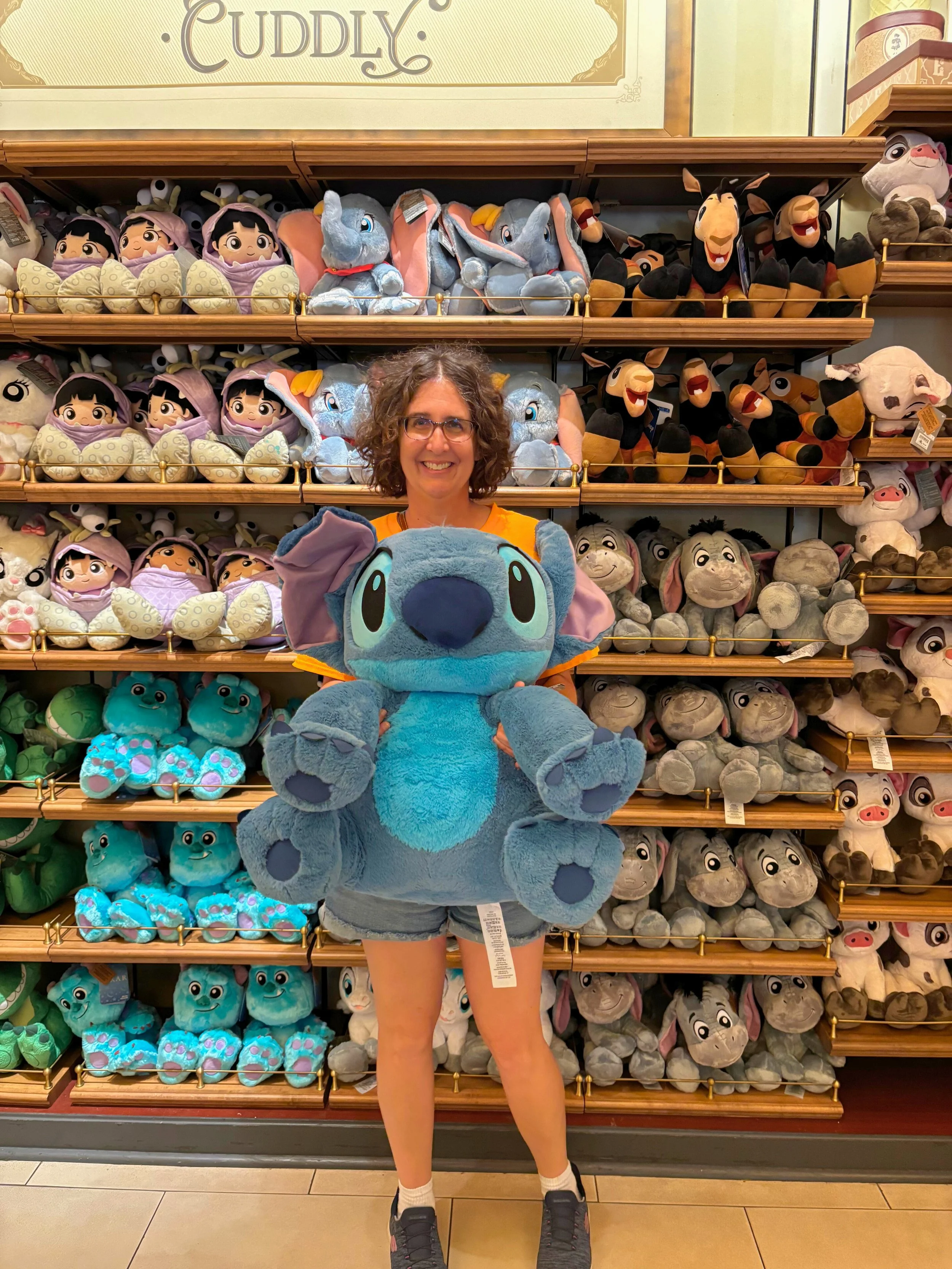 At Magic Kingdom, Maggie is standing in front of a selection of stuffed animals while holding a large Stich plush animal.