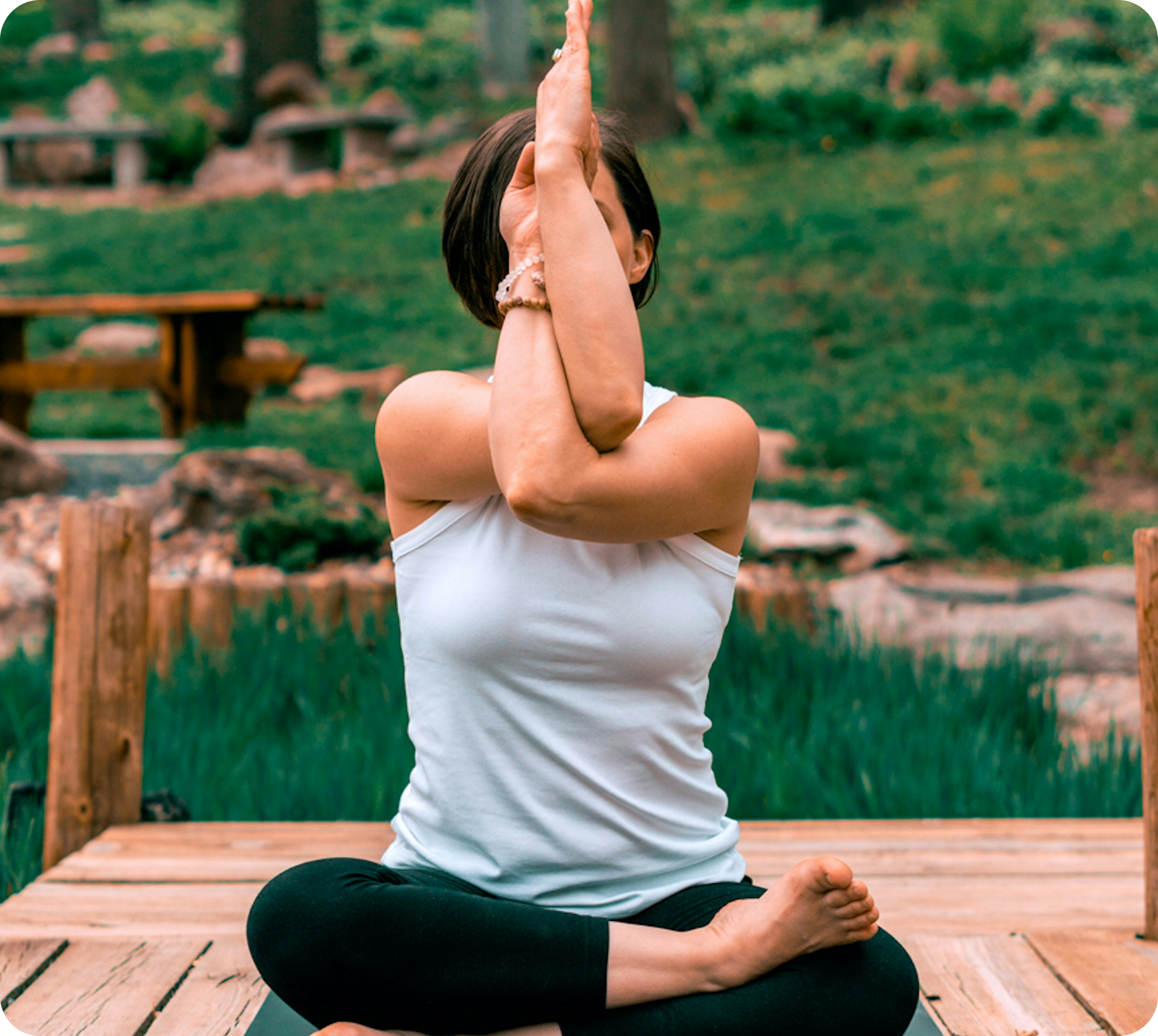 A woman practicing yoga outdoors on a wooden platform, sitting cross-legged with her hands pressed together in front of her face, her eyes closed, surrounded by greenery.