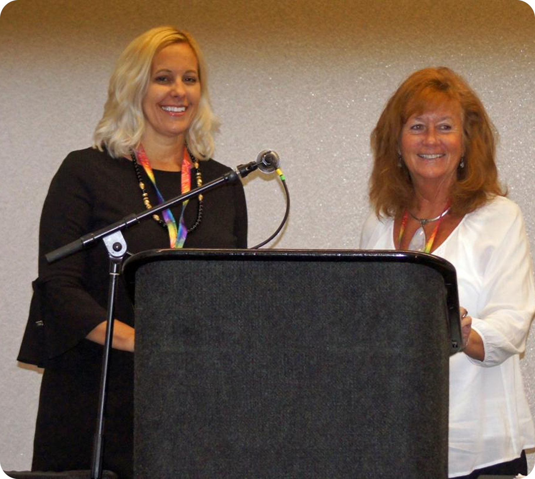 Trisha and Christy standing behind a podium with a microphone at an indoor event. Trisha has blonde hair and is wearing a black outfit. Christy has reddish hair and is wearing a white outfit.