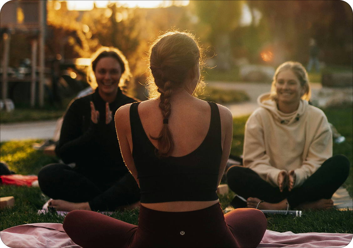 Three women sitting on the grass outdoors during sunset, engaging in a yoga or meditation session. One woman is seen from behind, sitting cross-legged with a braid, while the other two face her and smile, sitting cross-legged as well.