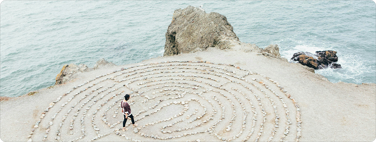 Person walking through a circular stone labyrinth on a coastal cliff overlooking the ocean.