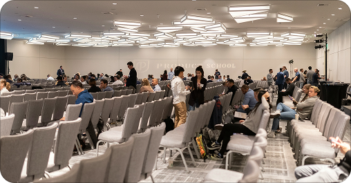 Conference room with rows of chairs and attendees seated, some standing and engaged in conversations, large screen at the front, ceiling with modern lighting fixtures.
