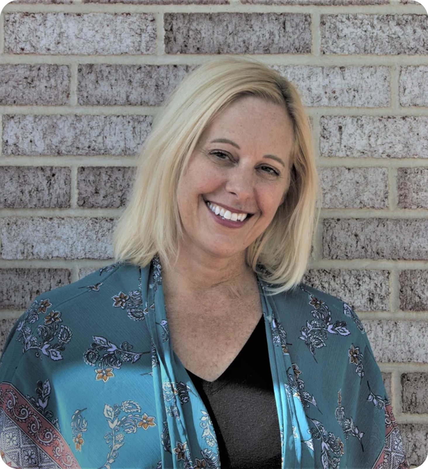 Trisha smiling with blonde hair standing in front of a brick wall, wearing a blue floral patterned shawl over a black top.