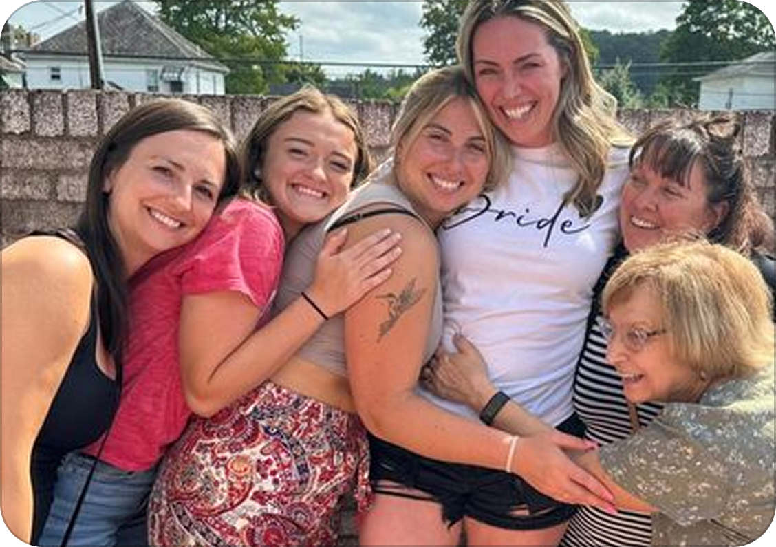 A group of six women smiling and hugging outdoors, with a brick wall and houses in the background.