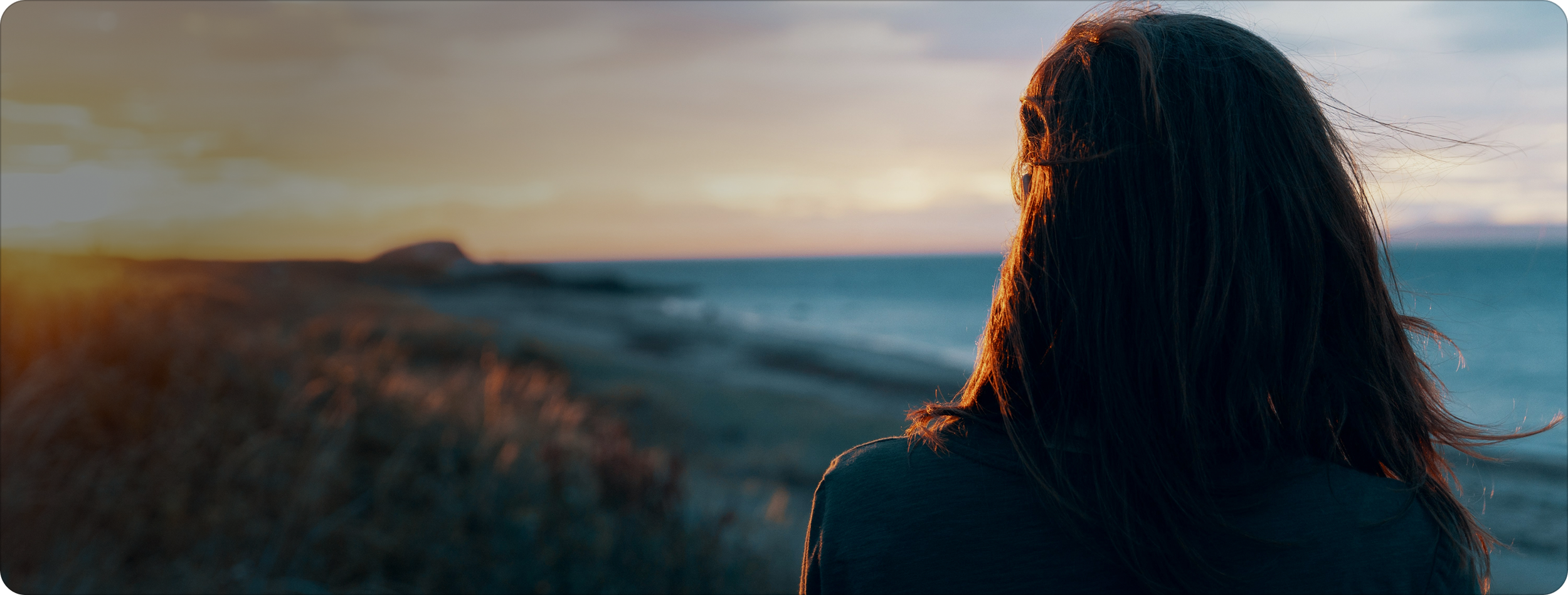 A woman with long hair facing the ocean at sunset, with the coastline and clouds in the background.
