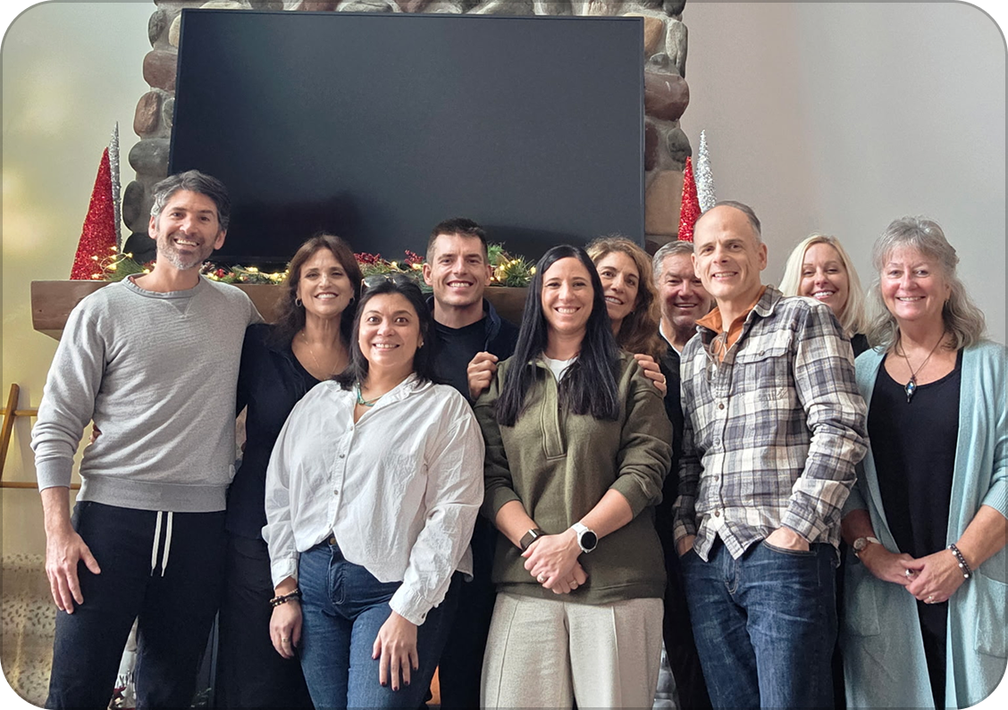 Group of ten people gathered together indoors, smiling, with a stone fireplace and decorated mantel in the background, suggesting a holiday celebration.