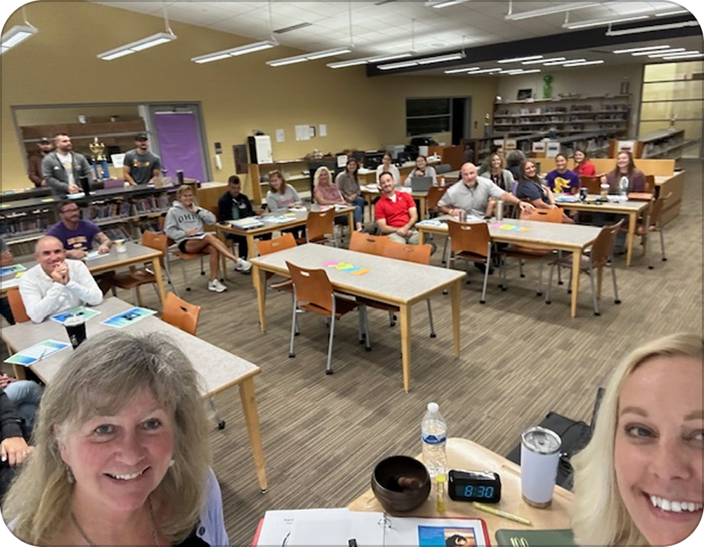 Group of people seated around tables in a library or classroom setting, smiling at the camera, with some standing behind the tables, and Trisha and Christy taking a selfie in the foreground.