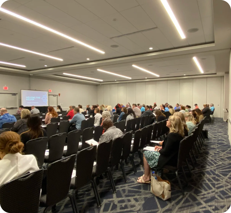 A large conference room filled with people seated in black chairs, watching a presentation on a projection screen that displays 'Helping the Helpline.' The room has bright ceiling lights and walls with quick access doors, and a patterned carpet.