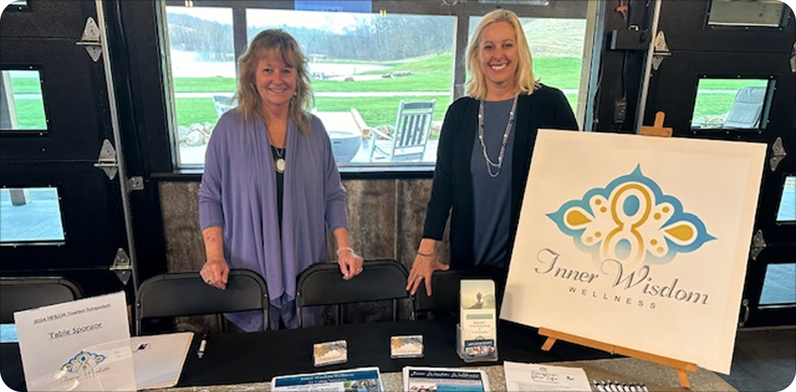 Trisha and Christy standing behind a table at an indoor event, with a large sign that reads 'Inner Wisdom Wellness'. The table displays informational materials and business cards. Large windows show an outdoor view of a lawn and a dock.