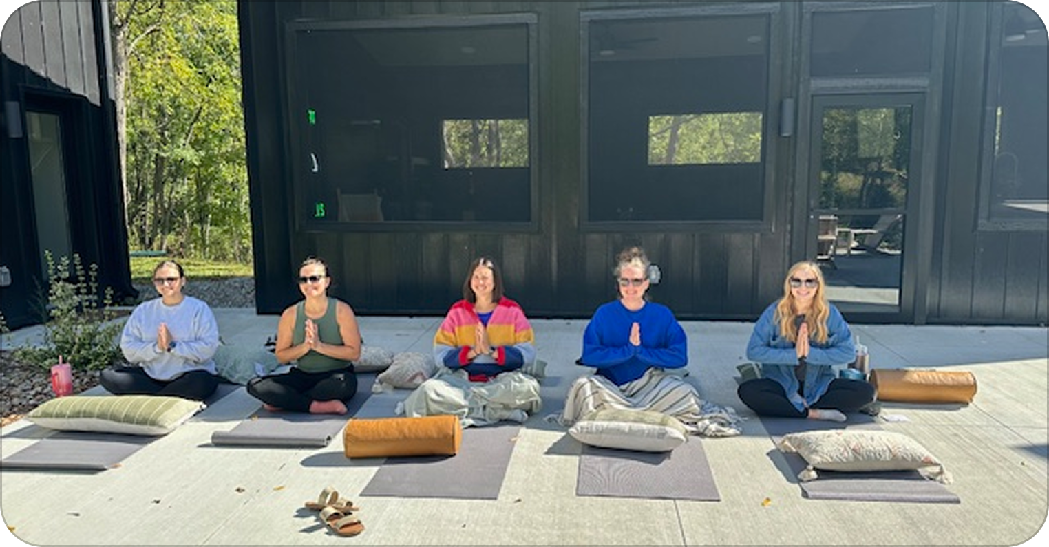 Five women practicing yoga outdoors, sitting on mats with pillows, in a village setting with trees in the background.