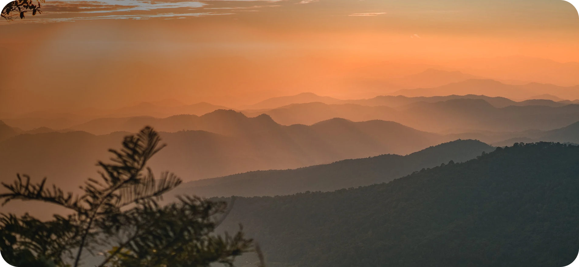 Multiple layers of mountain ridges at sunset with pink and orange sky, silhouetted trees in the foreground.