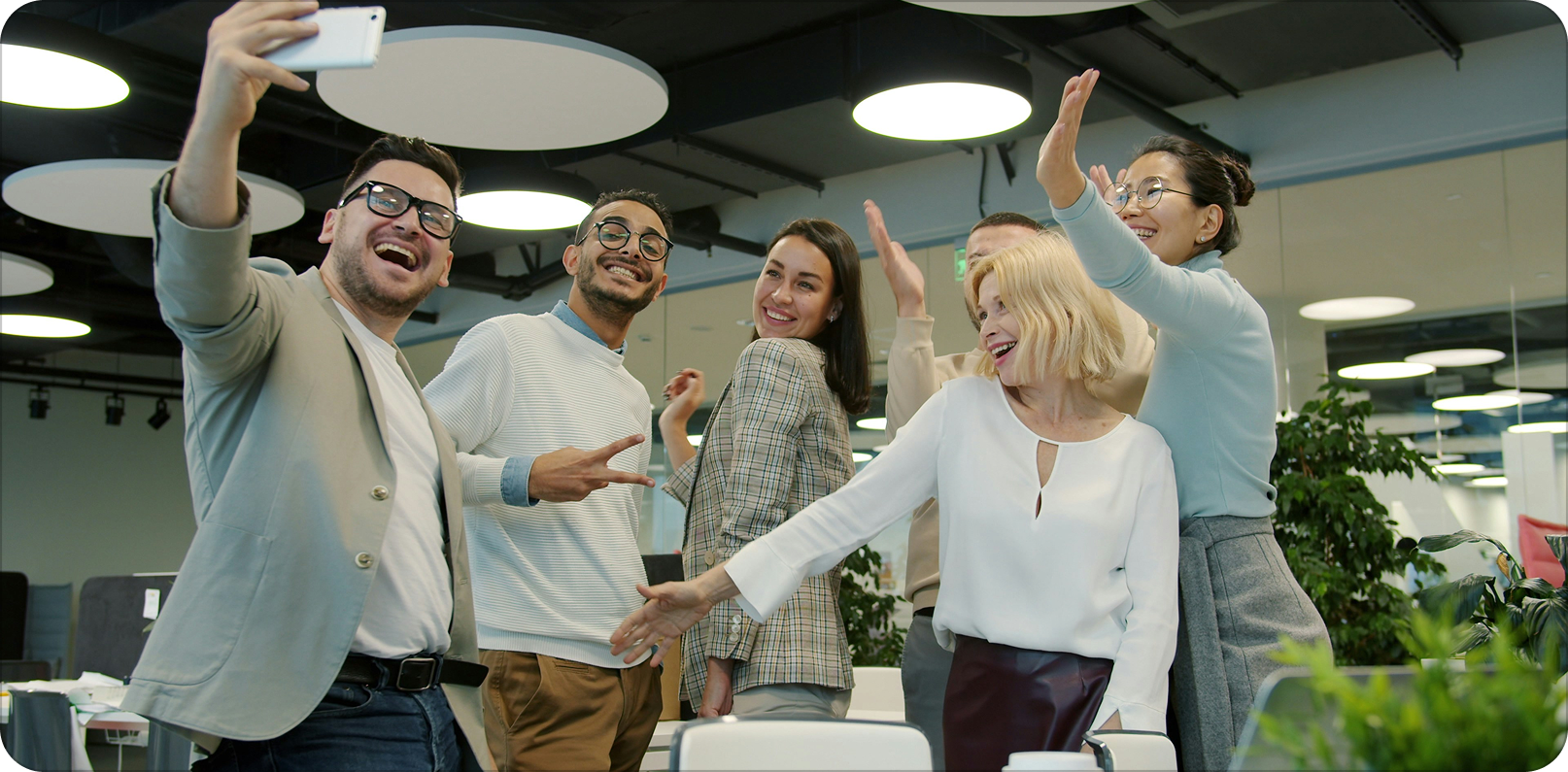 Group of six diverse coworkers smiling, high-fiving, and taking a selfie in an office.