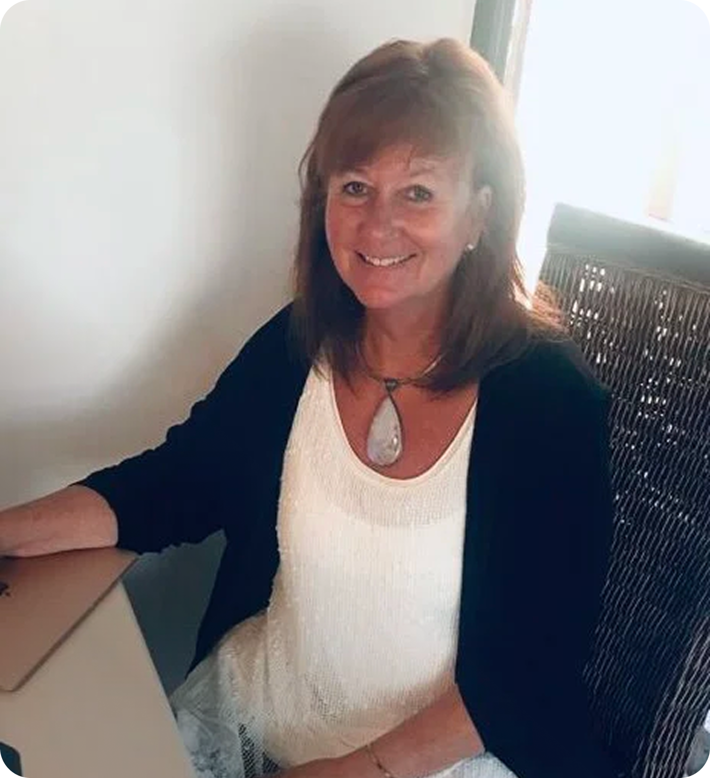 Christy with shoulder-length red hair, wearing a white top, black cardigan, and a large pendant necklace, smiling while sitting at a desk with a laptop.