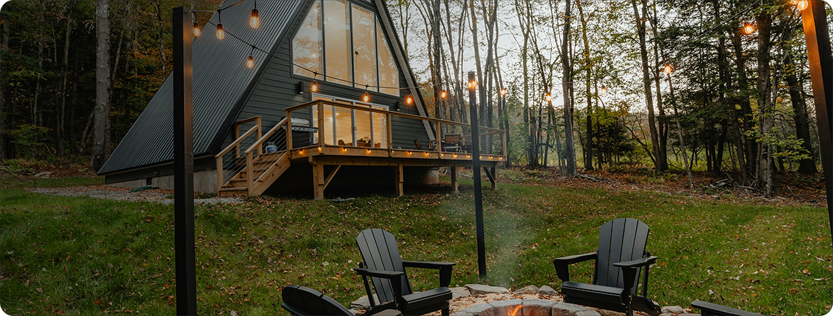 A cozy outdoor fire pit with black Adirondack chairs in a wooded backyard, outside a modern A-frame house decorated with string lights at dusk.