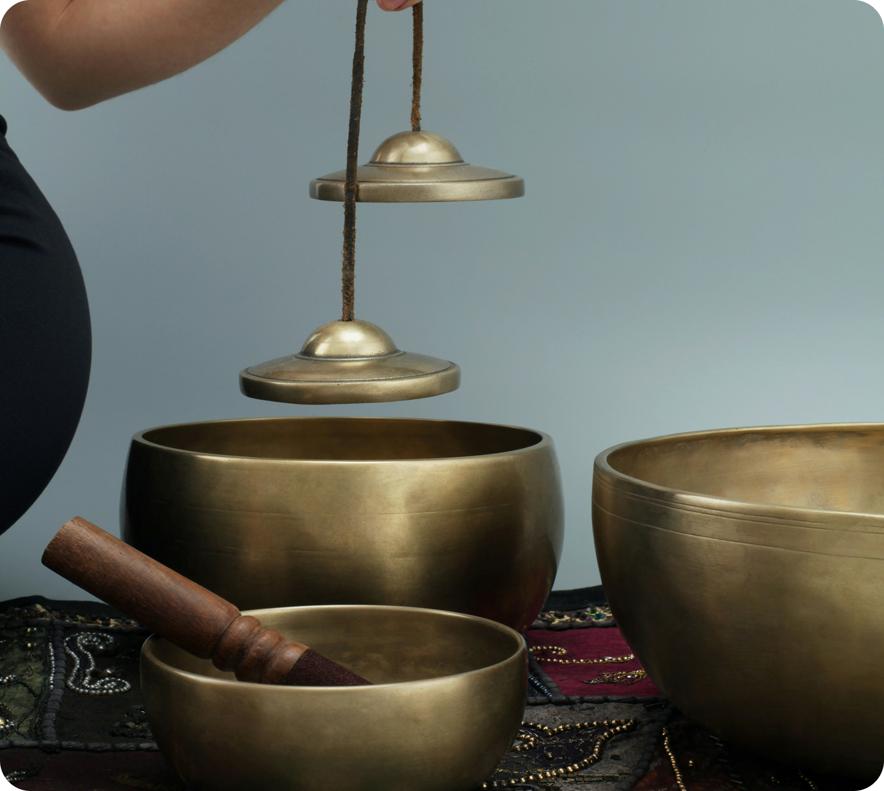 Three brass singing bowls of various sizes, one of which being struck with a wooden mallet, on an embroidered cloth.
