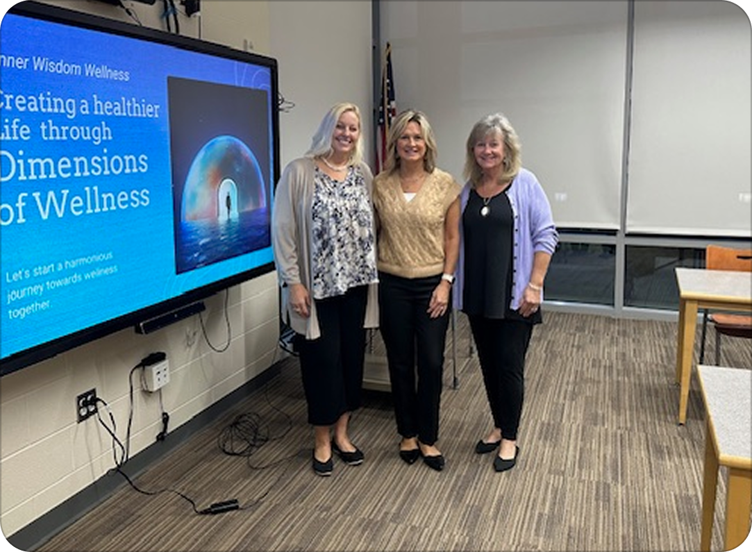 Three women standing in a room next to a large screen displaying a presentation slide titled 'Creating a healthier life through Dimensions of Wellness.' They are smiling at the camera and dressed in business casual attire.
