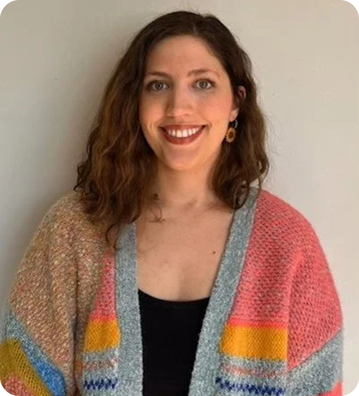 A woman with shoulder-length curly auburn hair, smiling, wearing a black top and a colorful knitted cardigan, standing against a plain light-colored wall.